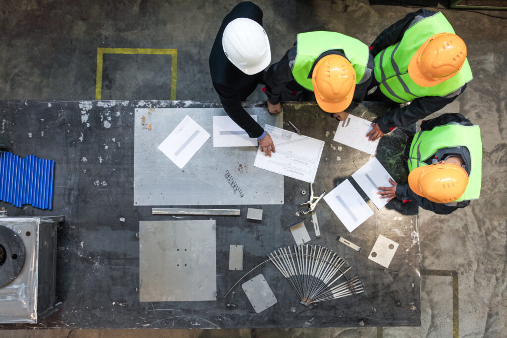Vier bouwvakkers in veiligheidskleding bespreken bouwtekeningen op een tafel in een magazijnomgeving.